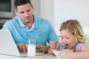 father with laptop looking at daughter having breakfast at table in house